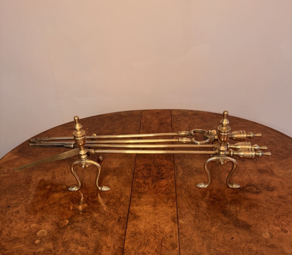 A set of three polished brass fire irons resting horizontally across a pair of matching brass fire dogs with urn-shaped finials. View V