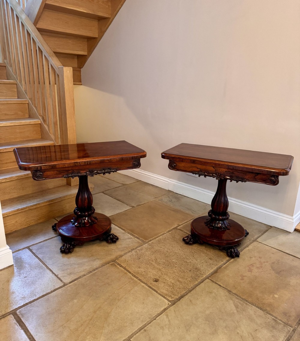 A pair of William IV rosewood card tables featuring swivel fold-over tops, carved friezes, central lobed pedestals, and circular plinth bases with lion paw feet.