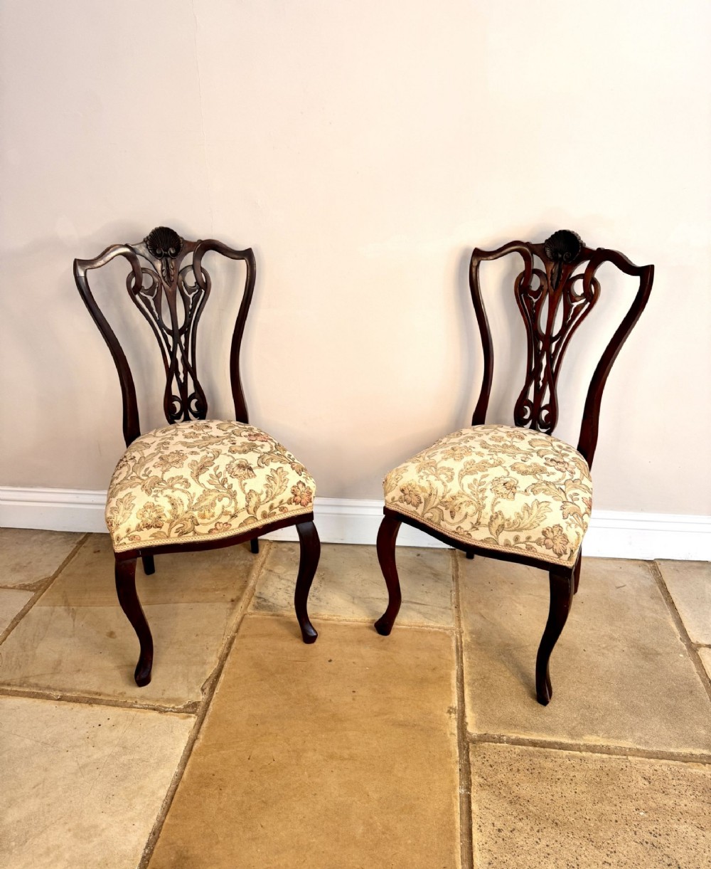 A pair of carved dark mahogany Victorian side chairs featuring ornate back splats with shell motifs, cabriole front legs, and cream floral pattern upholstery.