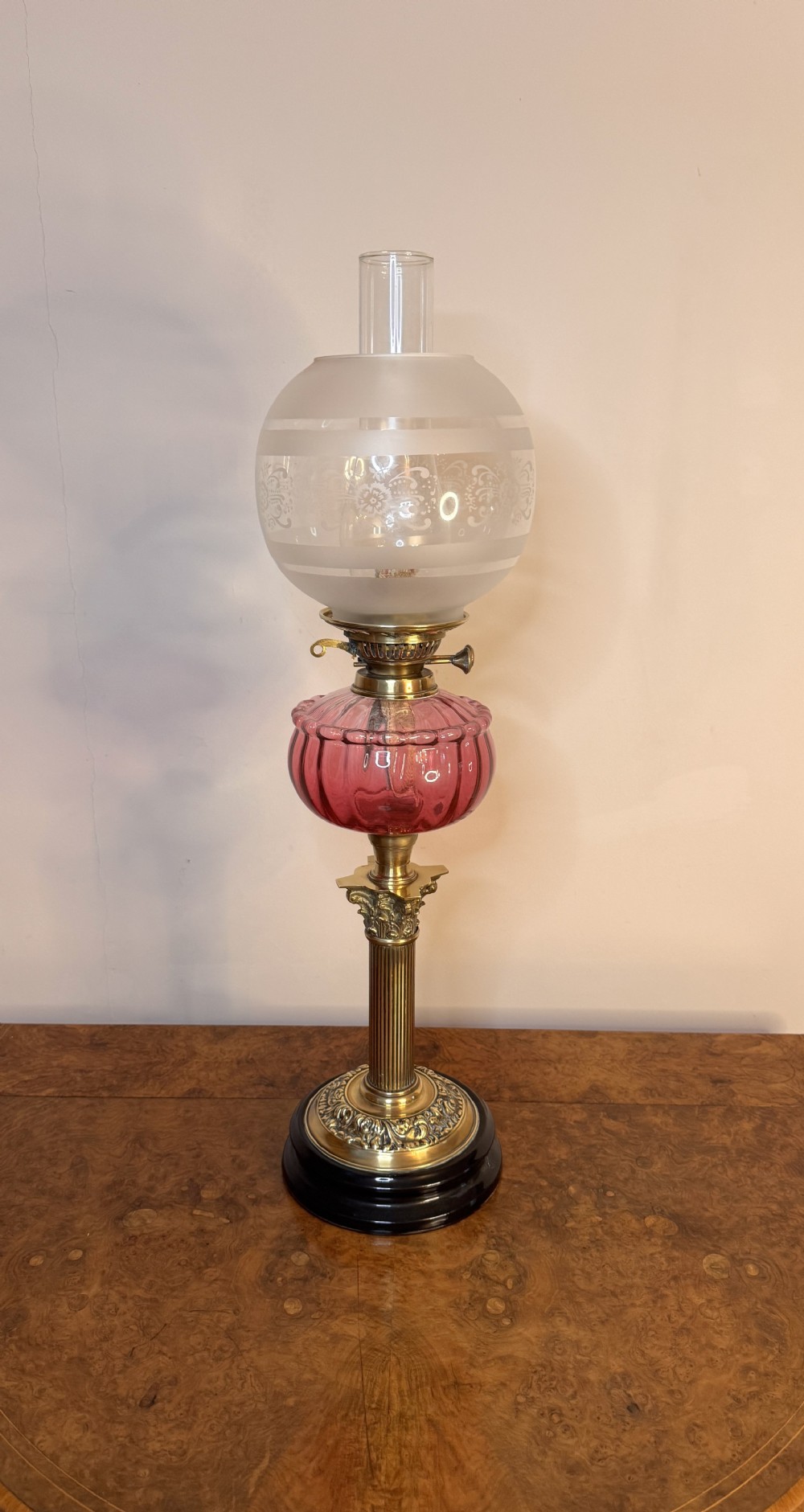 An antique Victorian oil lamp featuring a clear glass chimney, an etched frosted glass globe shade, a fluted cranberry glass reservoir, and a reeded brass Corinthian column base.