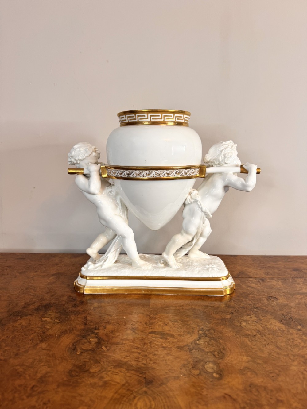A nineteenth-century white glazed porcelain centrepiece vessel supported on poles carried by two cherubs, detailed with gold gilded Greek key banding.