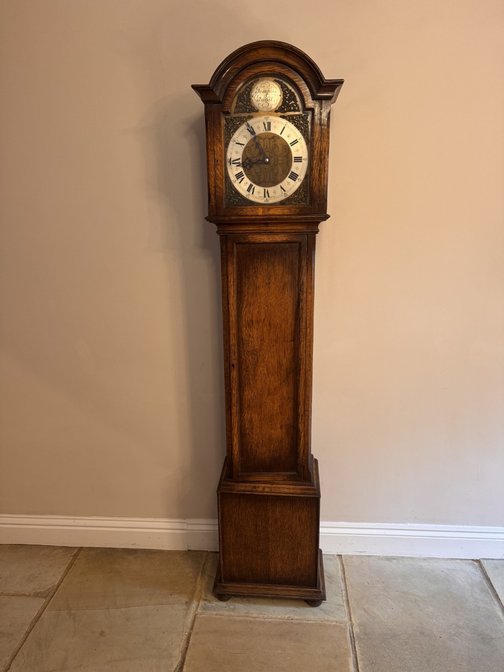 An antique oak longcase clock featuring an arched hood, a detailed brass dial with a Tempus Fugit crest, and a dark polished wooden casing.