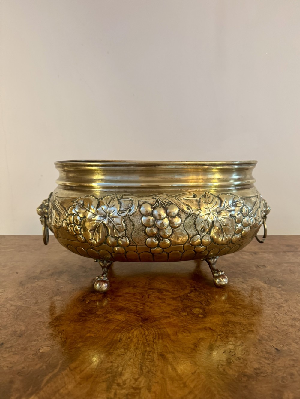 A round Victorian brass jardiniere planter featuring embossed grapevine relief, side lion mask ring handles, and four cast feet.