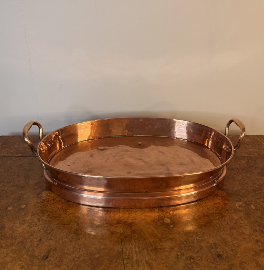 An oval Victorian copper pan featuring polished sides, a flat base, and two cast side handles secured with copper rivets.
