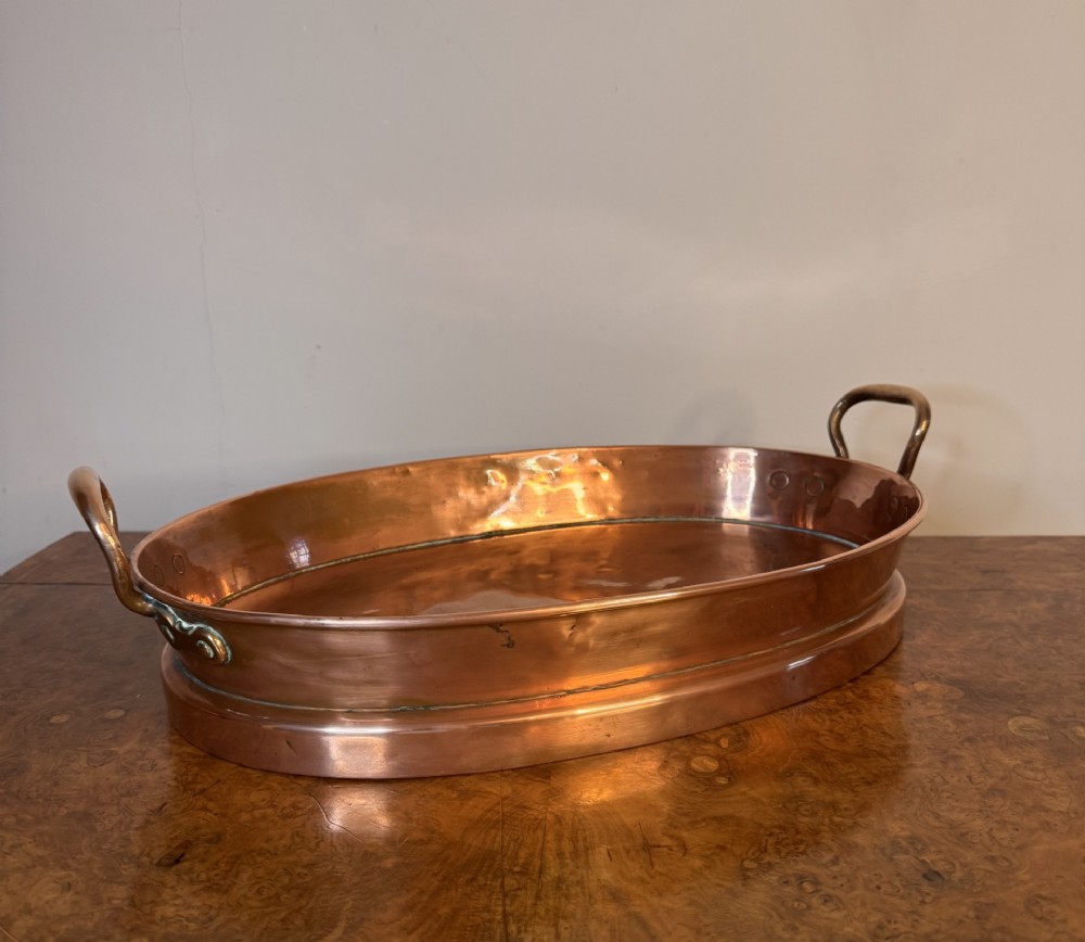 An oval Victorian copper pan featuring polished sides, a flat base, and two cast side handles secured with copper rivets. View VI
