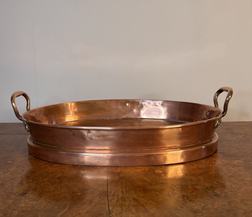 An oval Victorian copper pan featuring polished sides, a flat base, and two cast side handles secured with copper rivets. View VII