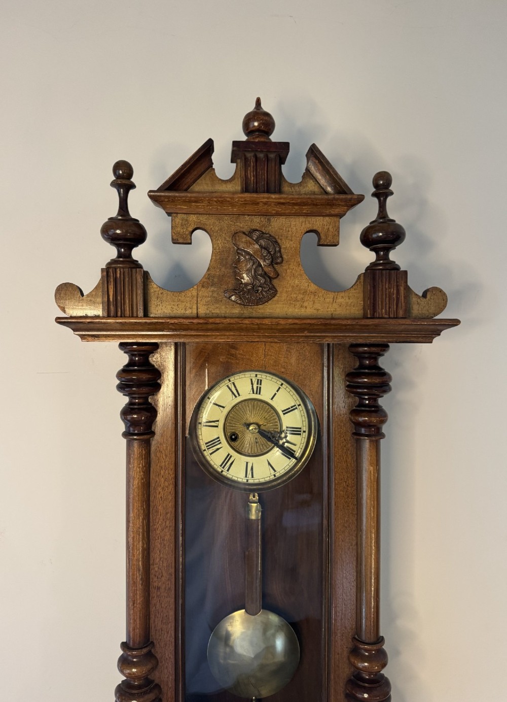An antique Victorian Vienna wall clock crafted in dark carved wood with turned pilasters, a decorative architectural pediment, glass door, Roman numeral dial, and visible brass pendulum. View II