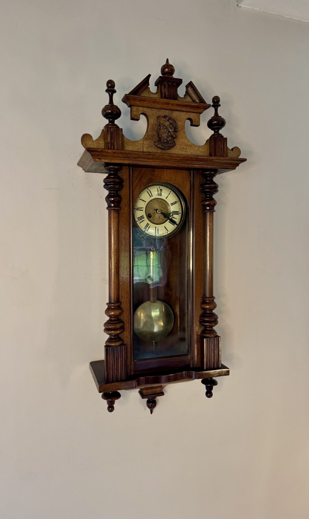 An antique Victorian Vienna wall clock crafted in dark carved wood with turned pilasters, a decorative architectural pediment, glass door, Roman numeral dial, and visible brass pendulum. View III