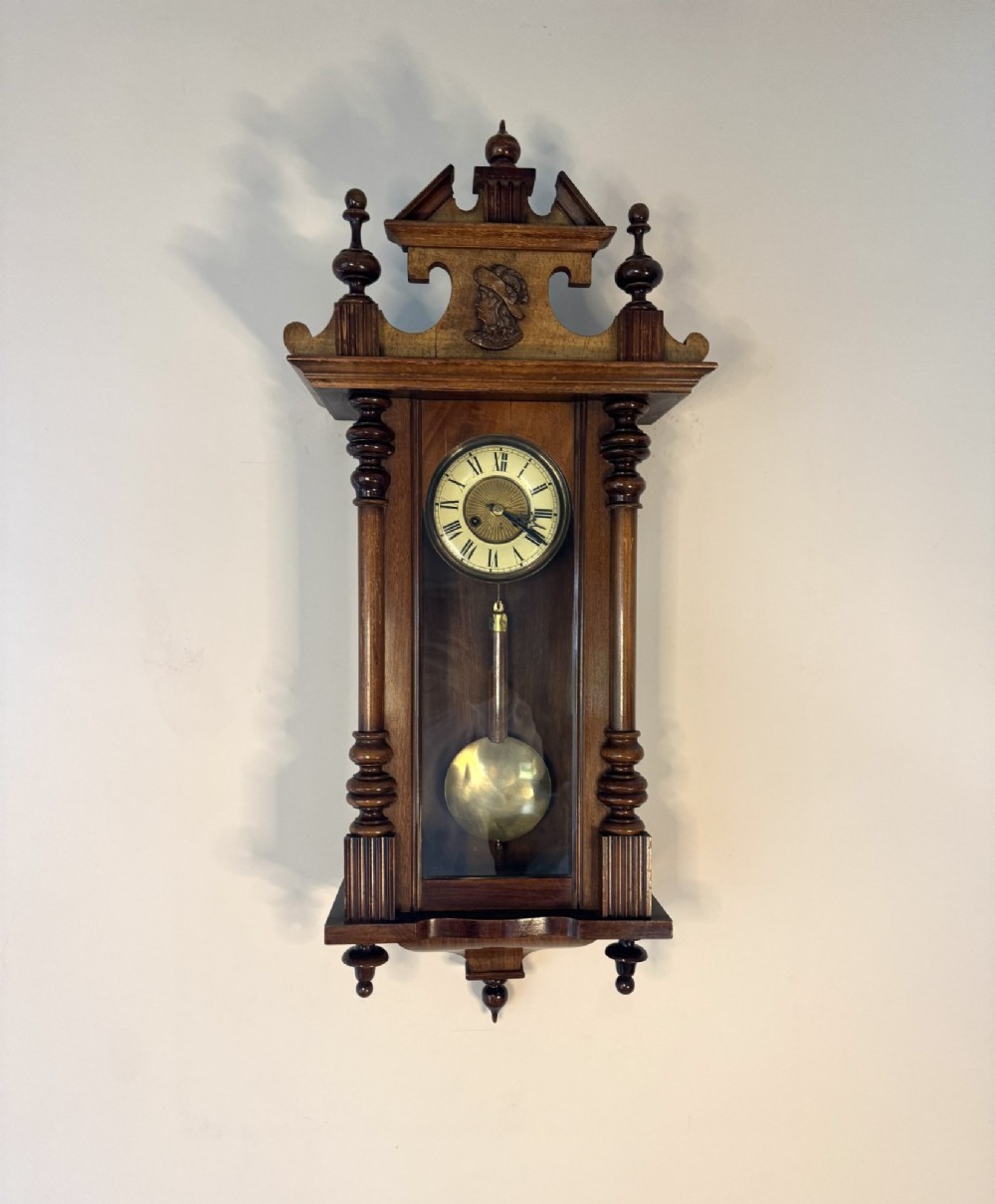 An antique Victorian Vienna wall clock crafted in dark carved wood with turned pilasters, a decorative architectural pediment, glass door, Roman numeral dial, and visible brass pendulum. View IV