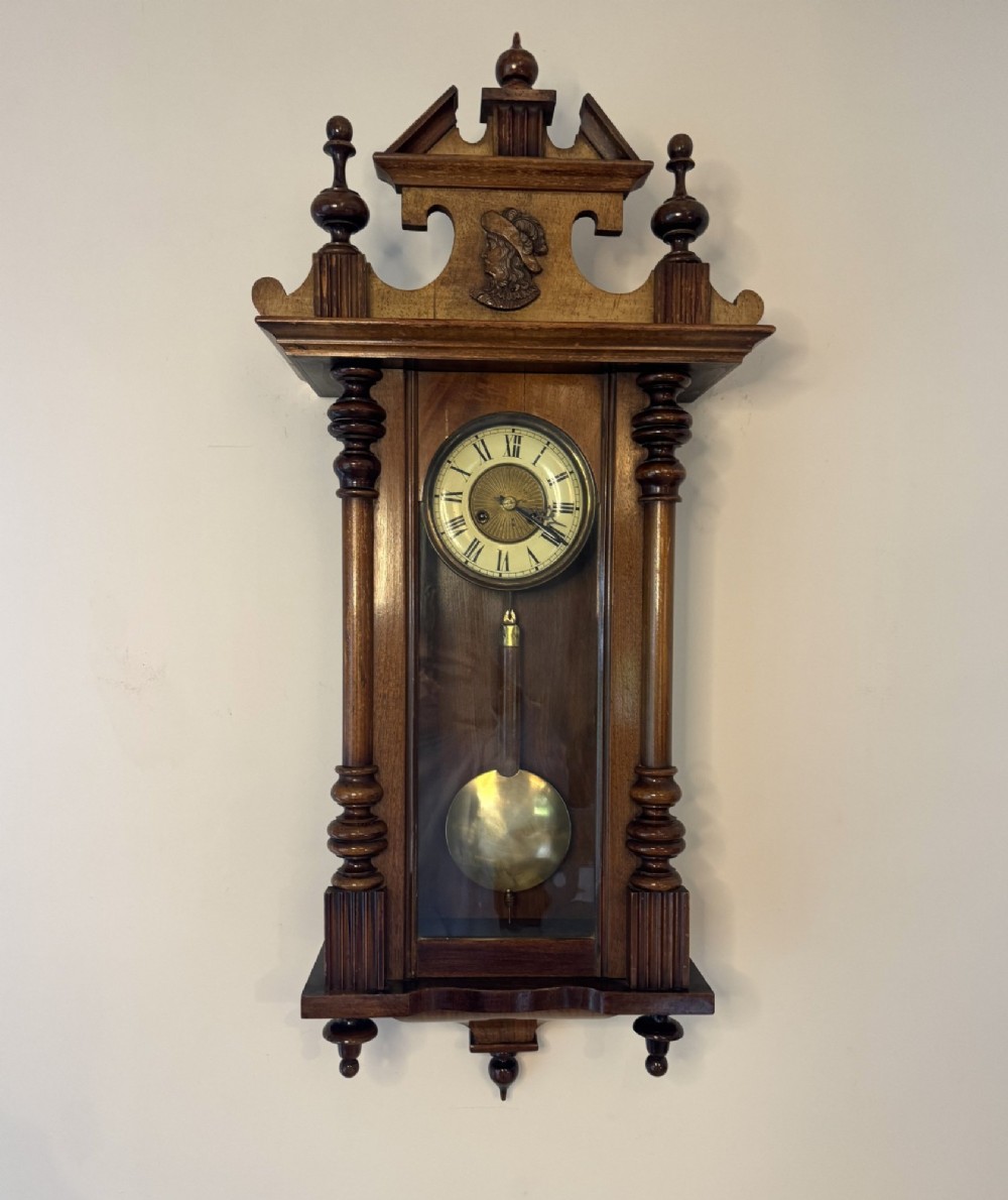 An antique Victorian Vienna wall clock crafted in dark carved wood with turned pilasters, a decorative architectural pediment, glass door, Roman numeral dial, and visible brass pendulum. View V