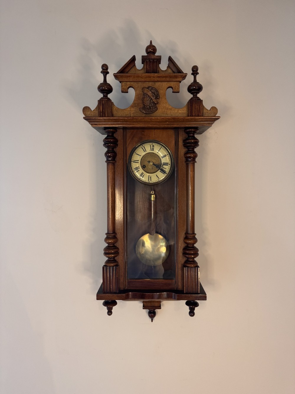 An antique Victorian Vienna wall clock crafted in dark carved wood with turned pilasters, a decorative architectural pediment, glass door, Roman numeral dial, and visible brass pendulum.