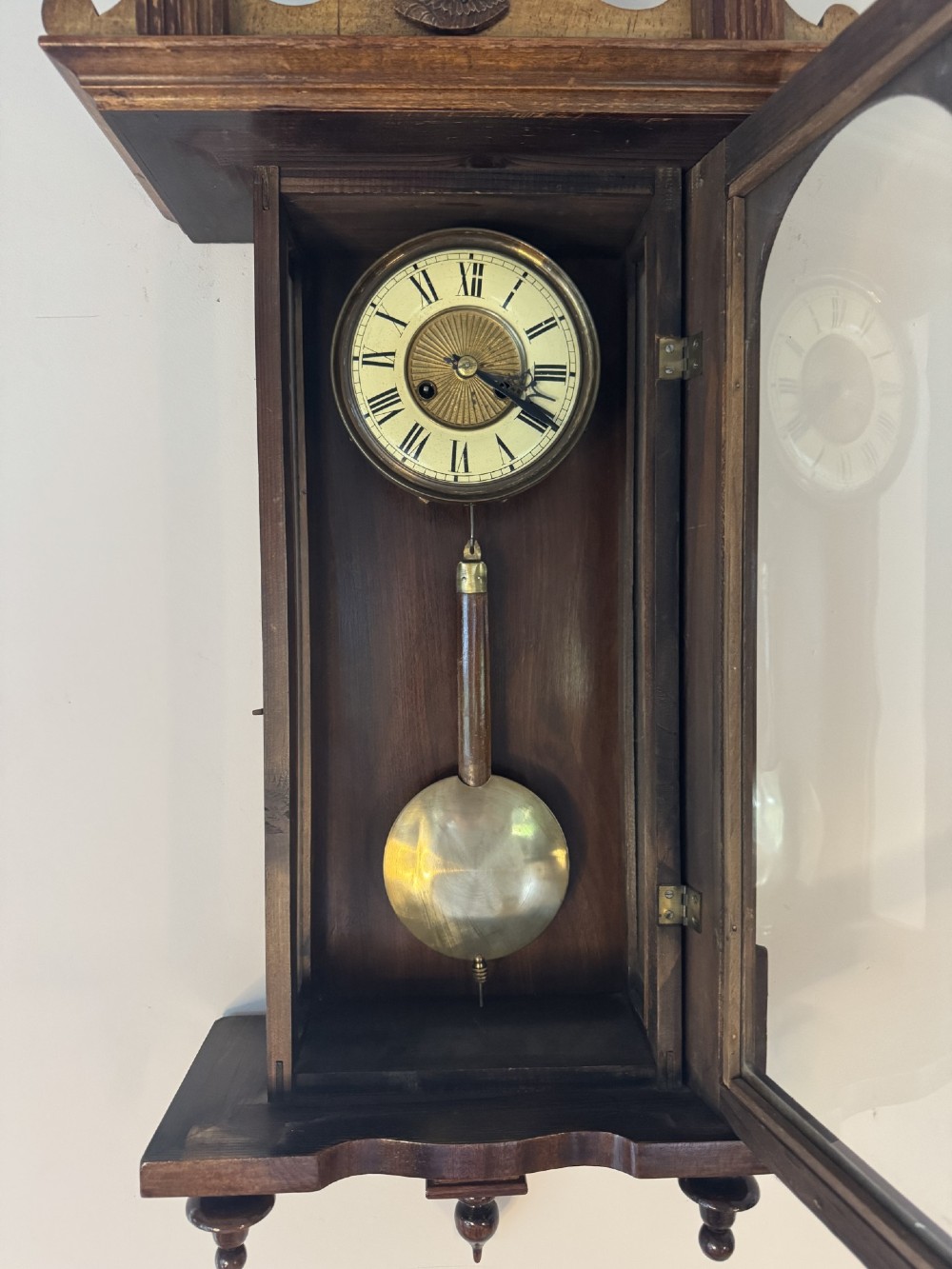 An antique Victorian Vienna wall clock crafted in dark carved wood with turned pilasters, a decorative architectural pediment, glass door, Roman numeral dial, and visible brass pendulum. View VI