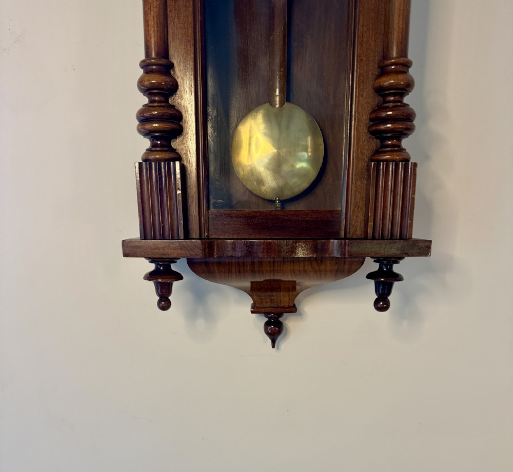 An antique Victorian Vienna wall clock crafted in dark carved wood with turned pilasters, a decorative architectural pediment, glass door, Roman numeral dial, and visible brass pendulum. View VII
