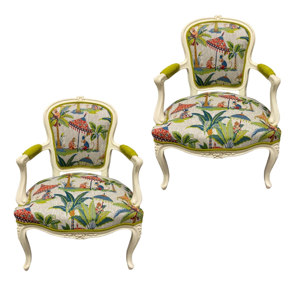 A pair of cream painted Louis XV style armchairs featuring carved cresting, cabriole legs, and upholstery with a monkey and palm tree pattern.