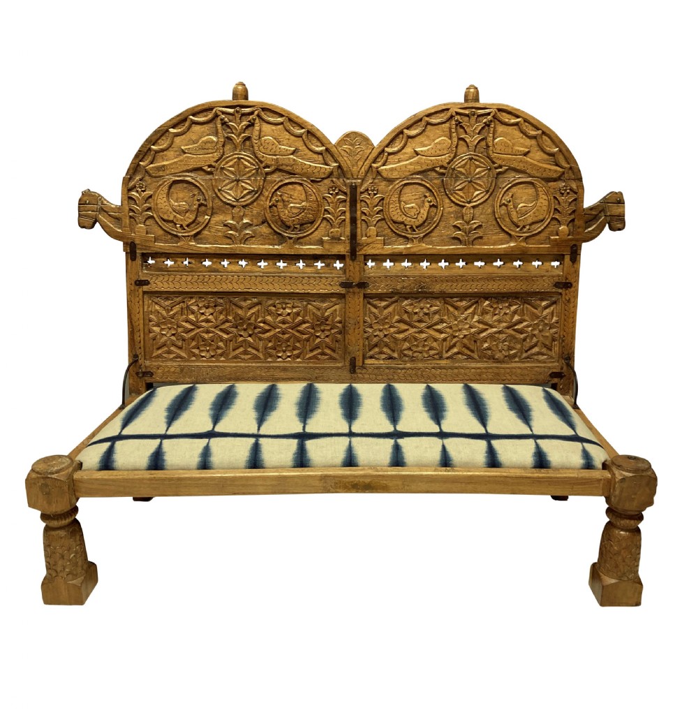 A low carved teak floor seat with arched backrest panels featuring carved bird motifs and a blue and white ikat patterned cushion.