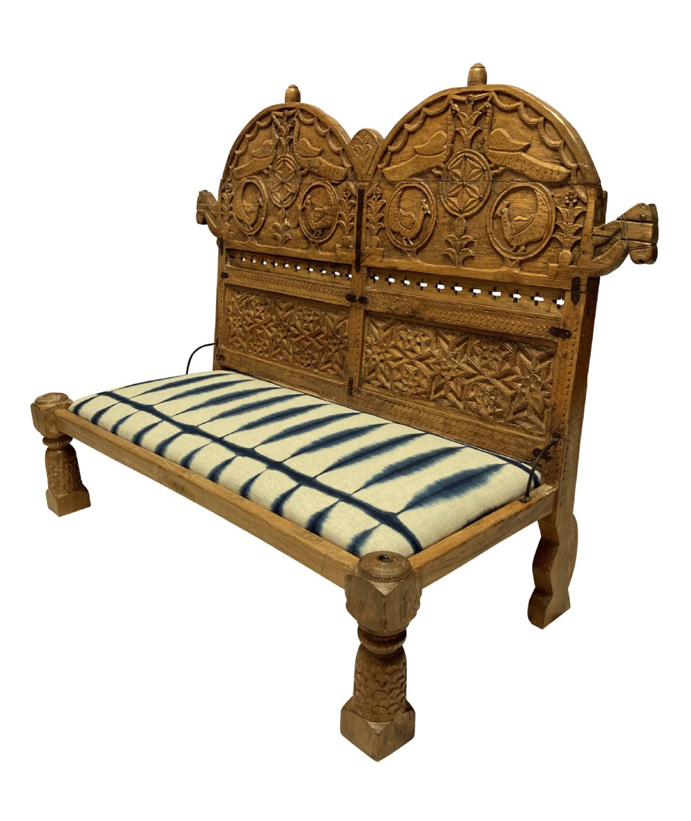 A low carved teak floor seat with arched backrest panels featuring carved bird motifs and a blue and white ikat patterned cushion. View IV