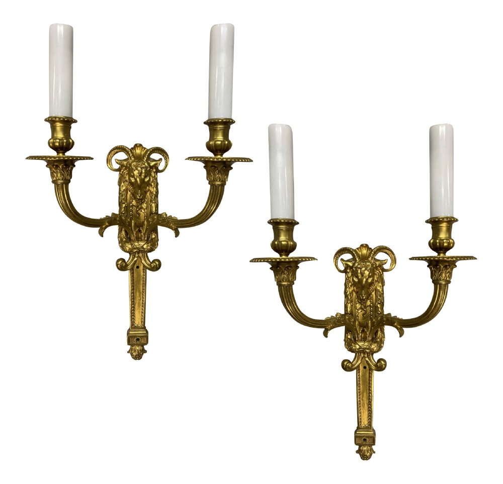 A pair of gilt brass Regency style twin-arm wall sconces featuring ram head central backplates and white candle sleeves.