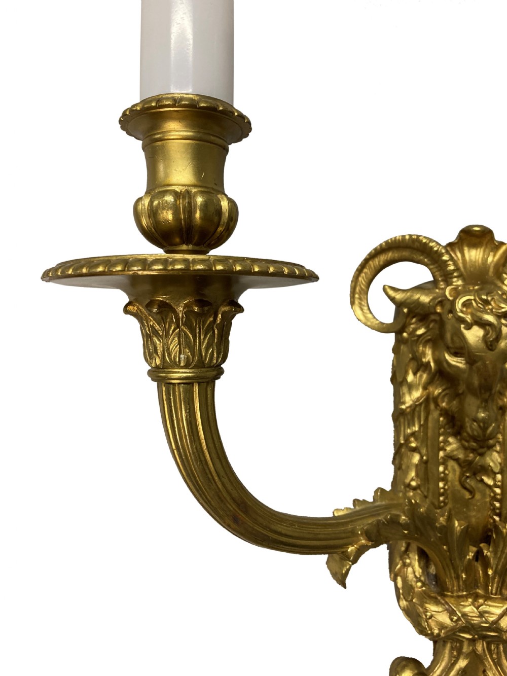 A pair of gilt brass Regency style twin-arm wall sconces featuring ram head central backplates and white candle sleeves. View V