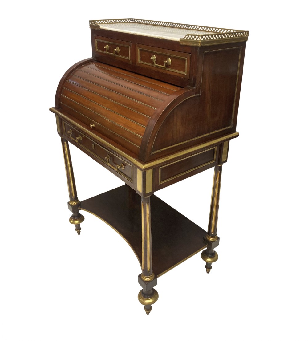 A small Directoire style mahogany cylinder desk with brass inlay trim, a marble top framed by a pierced brass gallery, slatted roll-top cylinder, drawer, and tapered legs.