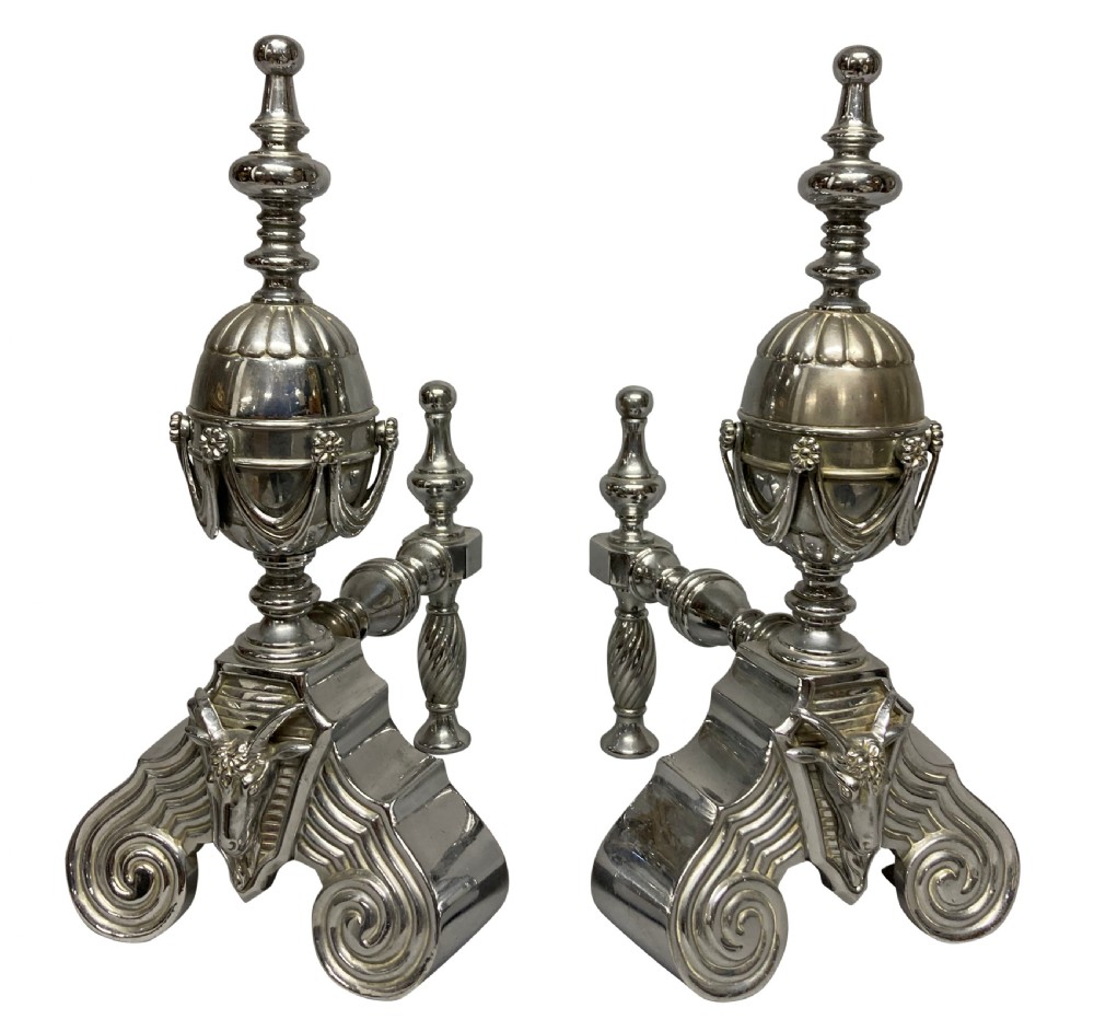 A pair of late 19th-century polished steel fire dogs featuring urn finials with swags, rams head reliefs, and scrolled bases.