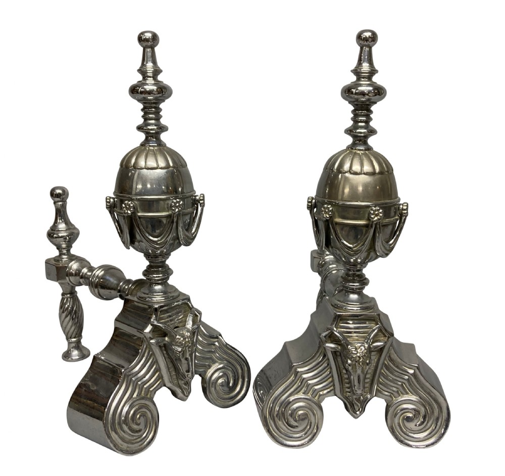 A pair of late 19th-century polished steel fire dogs featuring urn finials with swags, rams head reliefs, and scrolled bases. View II