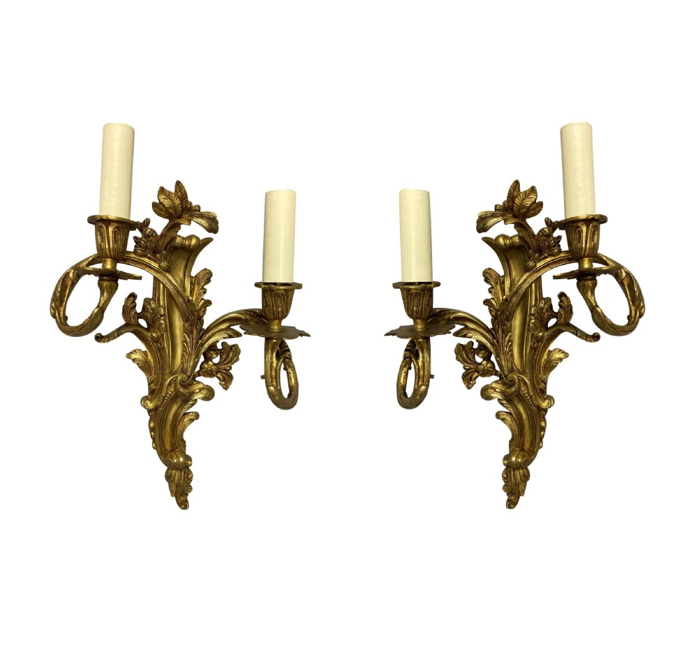 A pair of French Louis XV style gilt bronze two-light wall sconces featuring ornate asymmetrical acanthus foliage and candle holders.