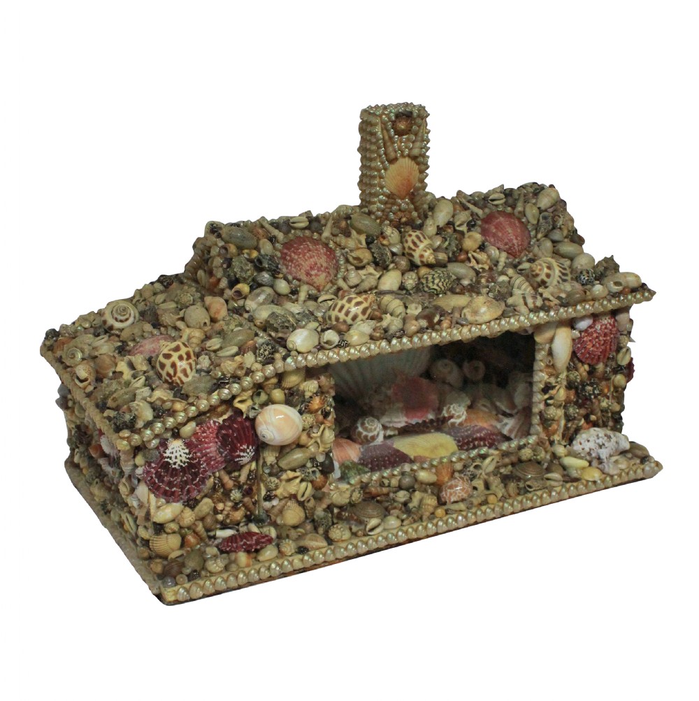 A miniature cottage-shaped decorative object encrusted with various small natural seashells and featuring a central open recess and a shell-covered chimney.