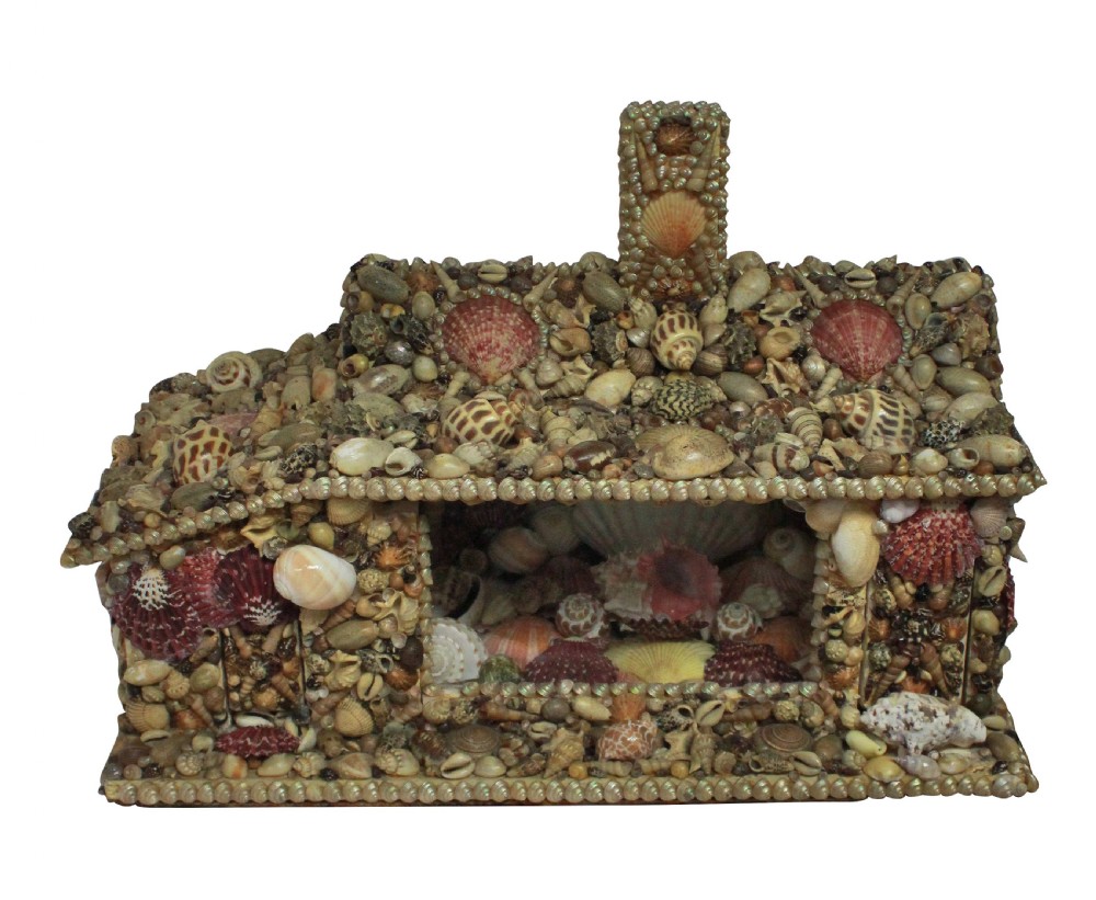 A miniature cottage-shaped decorative object encrusted with various small natural seashells and featuring a central open recess and a shell-covered chimney. View V