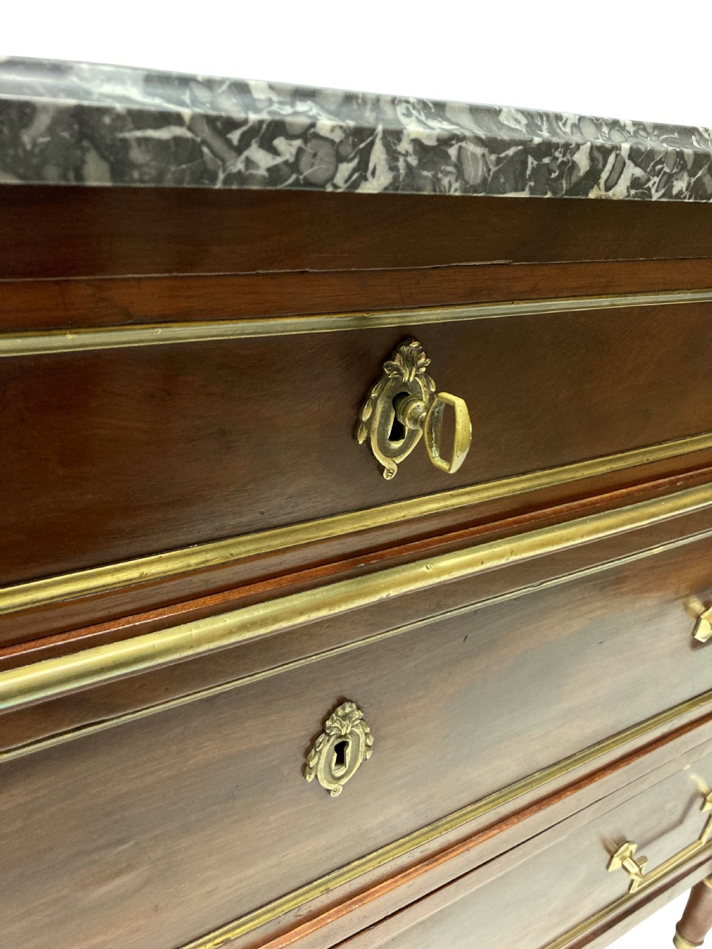 A French Directoire mahogany commode featuring three long drawers with brass mounts, fluted corner pilasters, and a grey marble top. View X