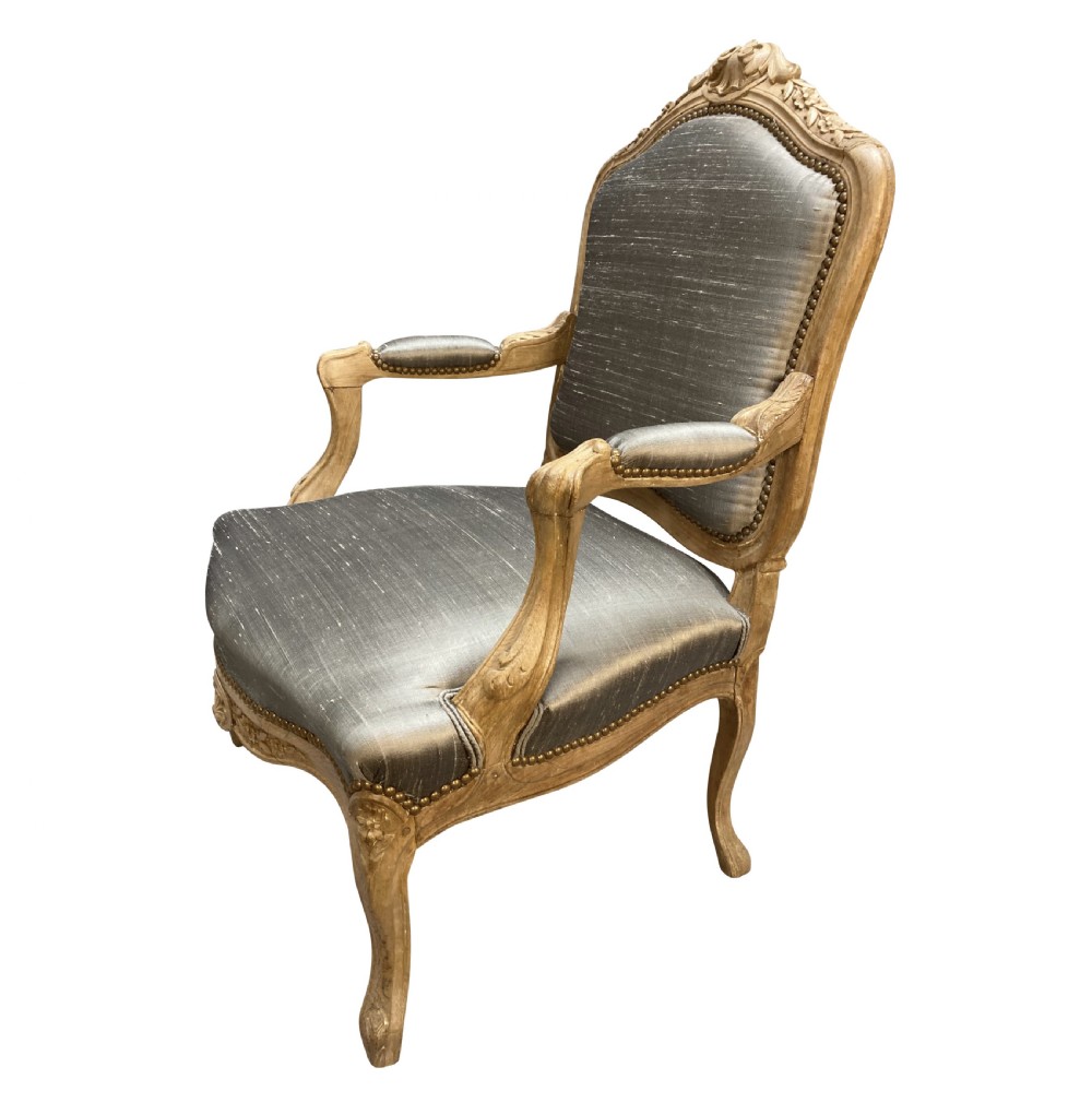 A Louis XV style armchair featuring a carved bleached fruitwood frame with cabriole legs and grey silk upholstery secured with brass studs.