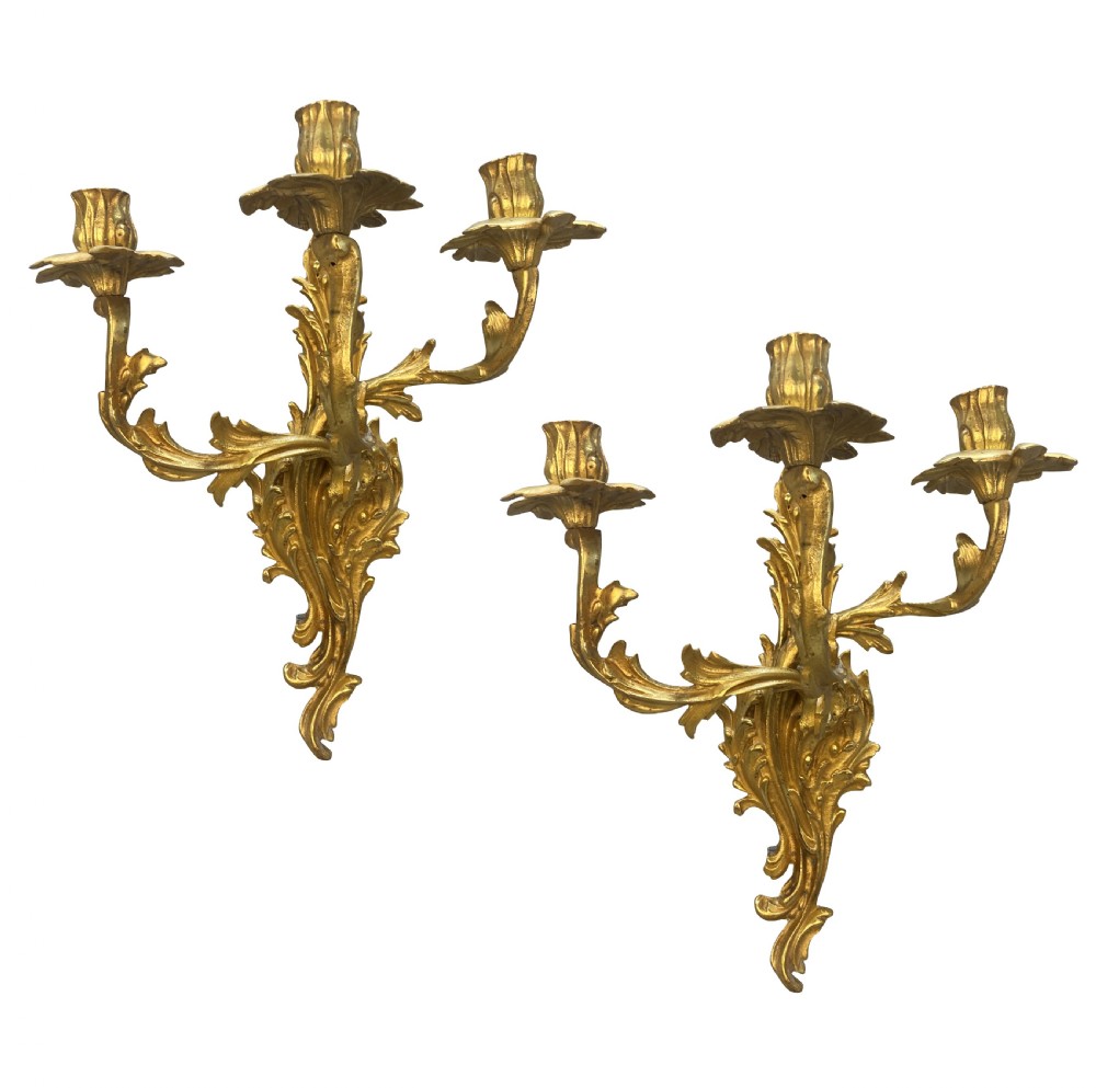A pair of Louis XV style gilt bronze three-arm wall sconces featuring scrolling foliage motifs and leaf-shaped drip pans.