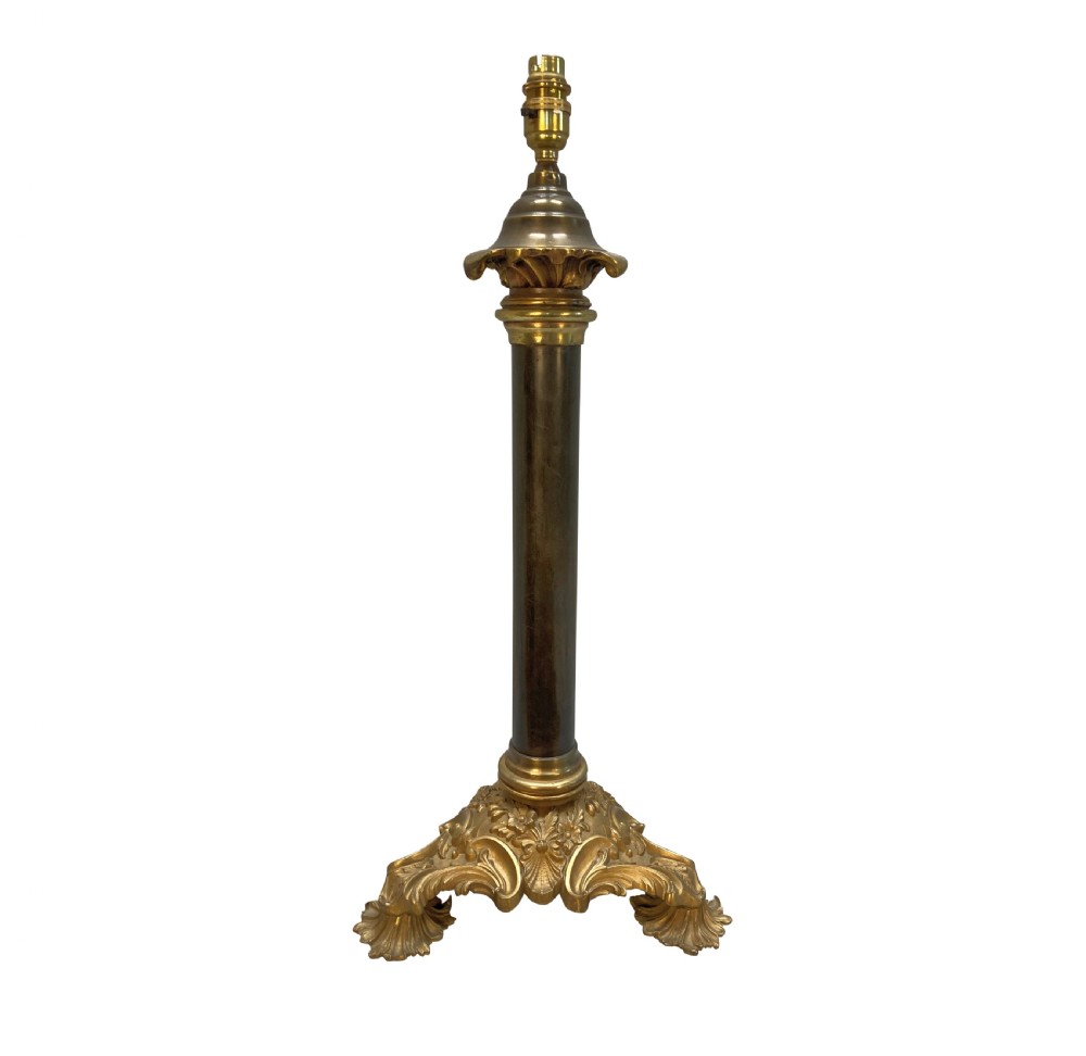 An English table lamp base featuring a dark patinated bronze cylindrical column mounted on a gilt ormolu triform base with scrolled feet and topped with an acanthus leaf capital.
