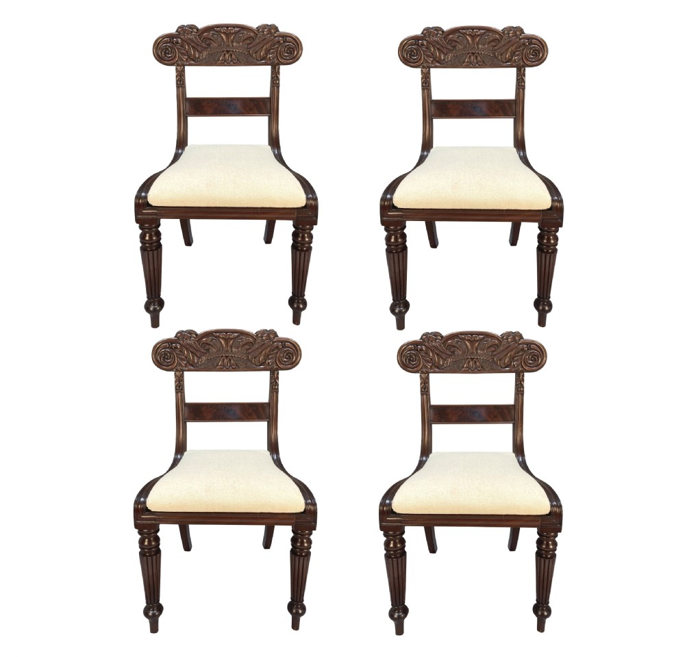 A set of four William IV Anglo-Indian dark wood dining chairs featuring carved scroll top rails, fluted turned front legs, and cream fabric seats.
