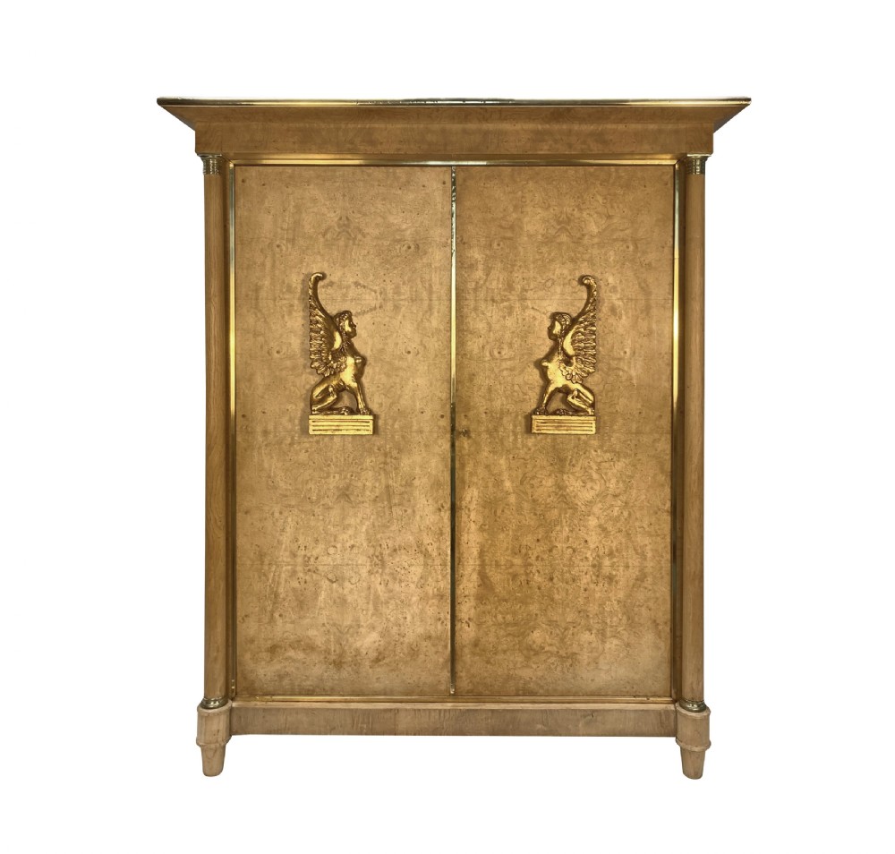 A large two-door Swedish Empire Revival cupboard crafted in light birds eye maple wood featuring gilt winged sphinx mounts and brass trim.