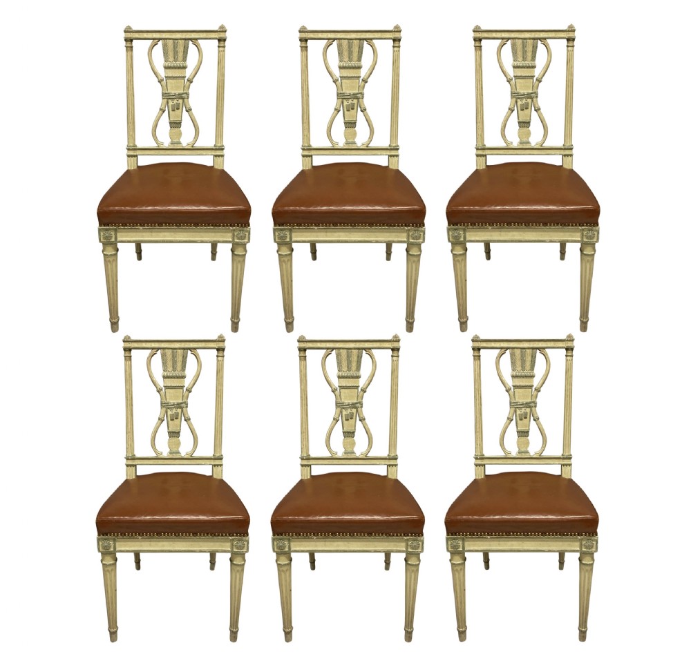 A set of six Louis XVI style cream painted side chairs featuring open carved backs with urn motifs and brown leather upholstered seats with stud detailing.