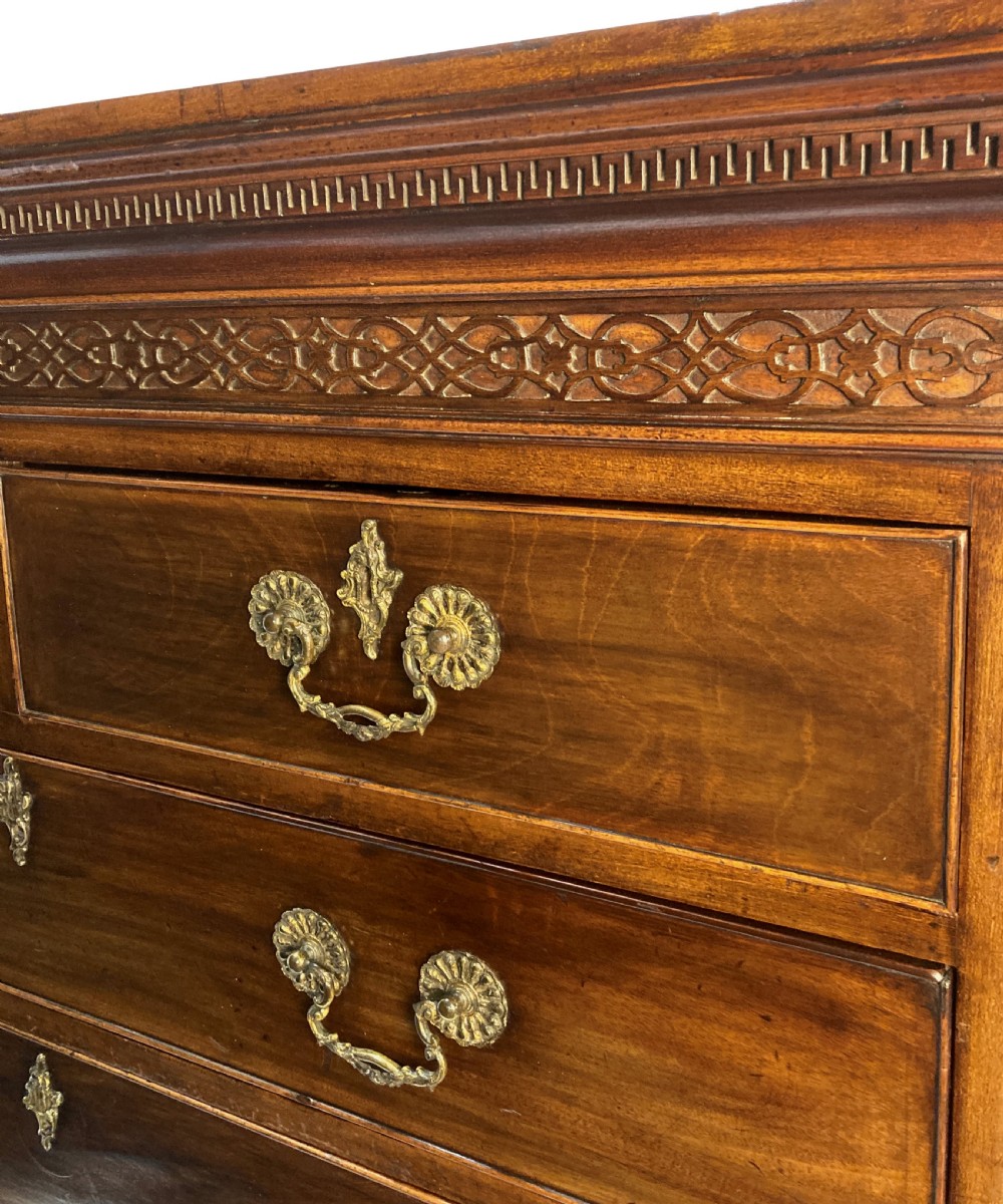 An English Chippendale style mahogany chest of drawers featuring ornate brass handles, a carved fretwork frieze, and bracket feet. View VI