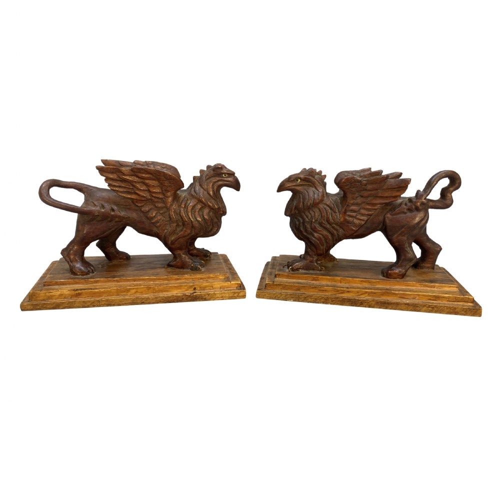 A pair of hand-carved wooden griffin statues facing each other, each mounted on a stepped rectangular plinth.