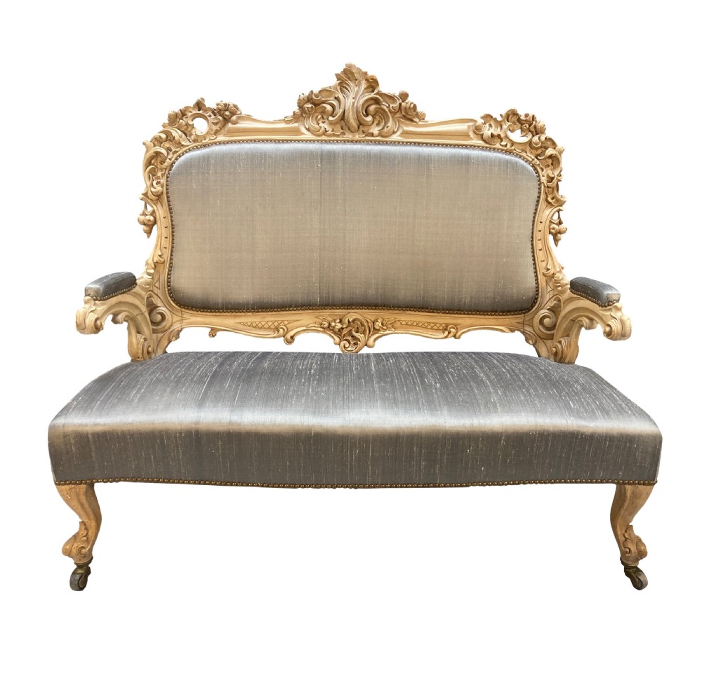 An early nineteenth-century German Rococo style upholstered settee with an elaborately carved crest rail, cabriole legs on castors, and silver-grey silk fabric seat and back.