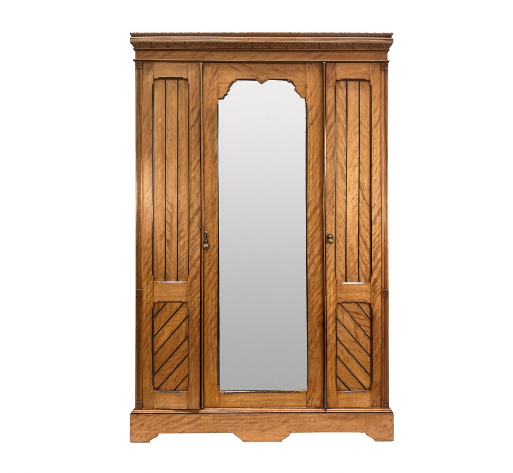 An Aesthetic Movement satin birch wardrobe featuring a central mirrored door flanked by geometric panelled side doors and a carved frieze.