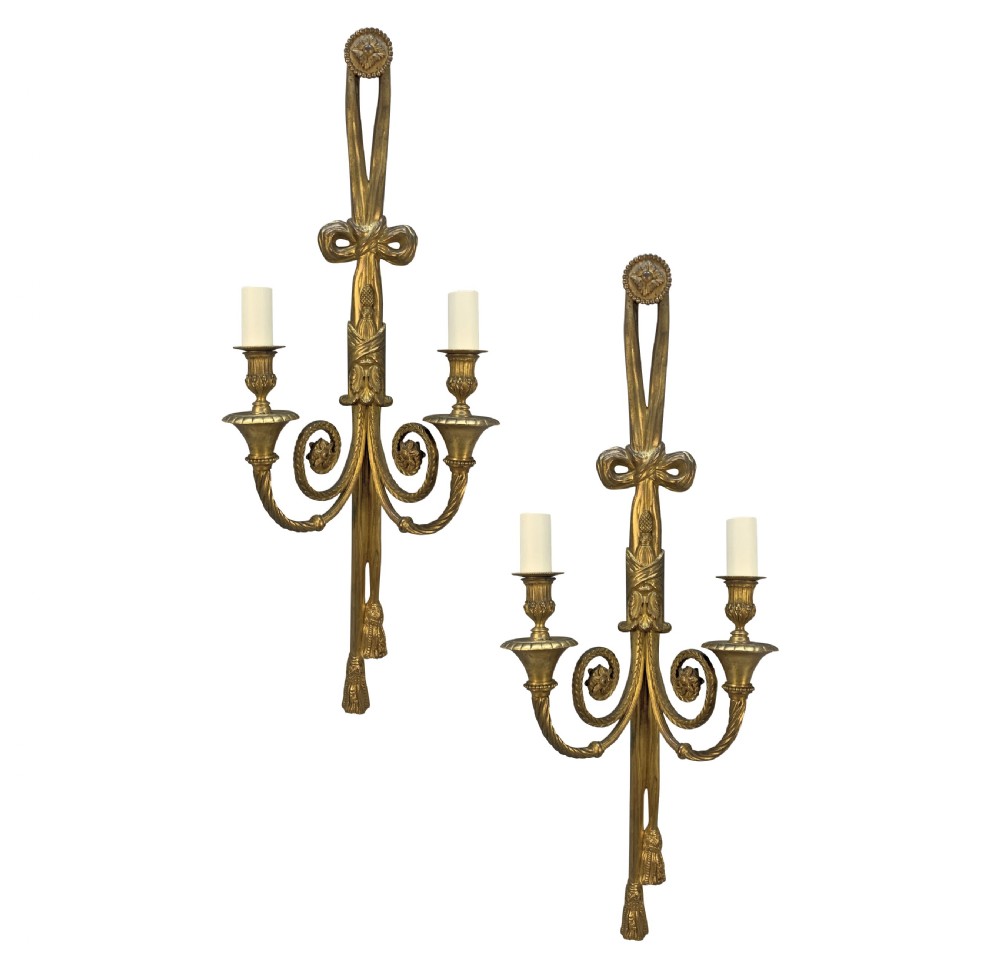 A pair of gilt bronze two-arm wall sconces featuring ribbon bow crests, tassel drops, and candle sockets.