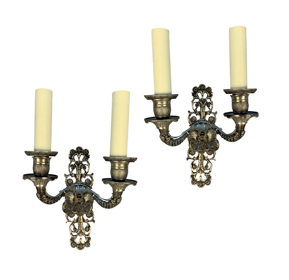 A pair of silver-plated French Empire style two-light wall sconces with ornate foliate backplates and cream candle sleeves.