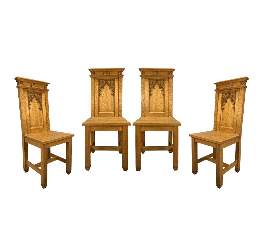 A set of four solid English oak hall chairs featuring carved Gothic arch panels on the backrests, flat solid seats, and straight legs with H-stretchers.