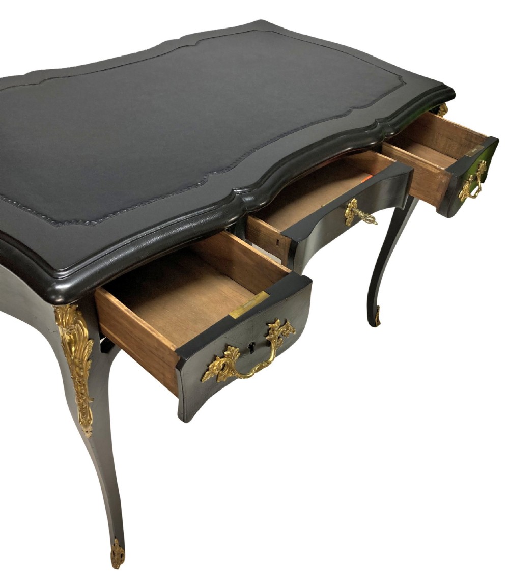A black lacquered Louis XV style writing table featuring a serpentine top with a leather writing surface, decorative gilt bronze mounts, three frieze drawers, and cabriole legs with gilded sabots. View IX