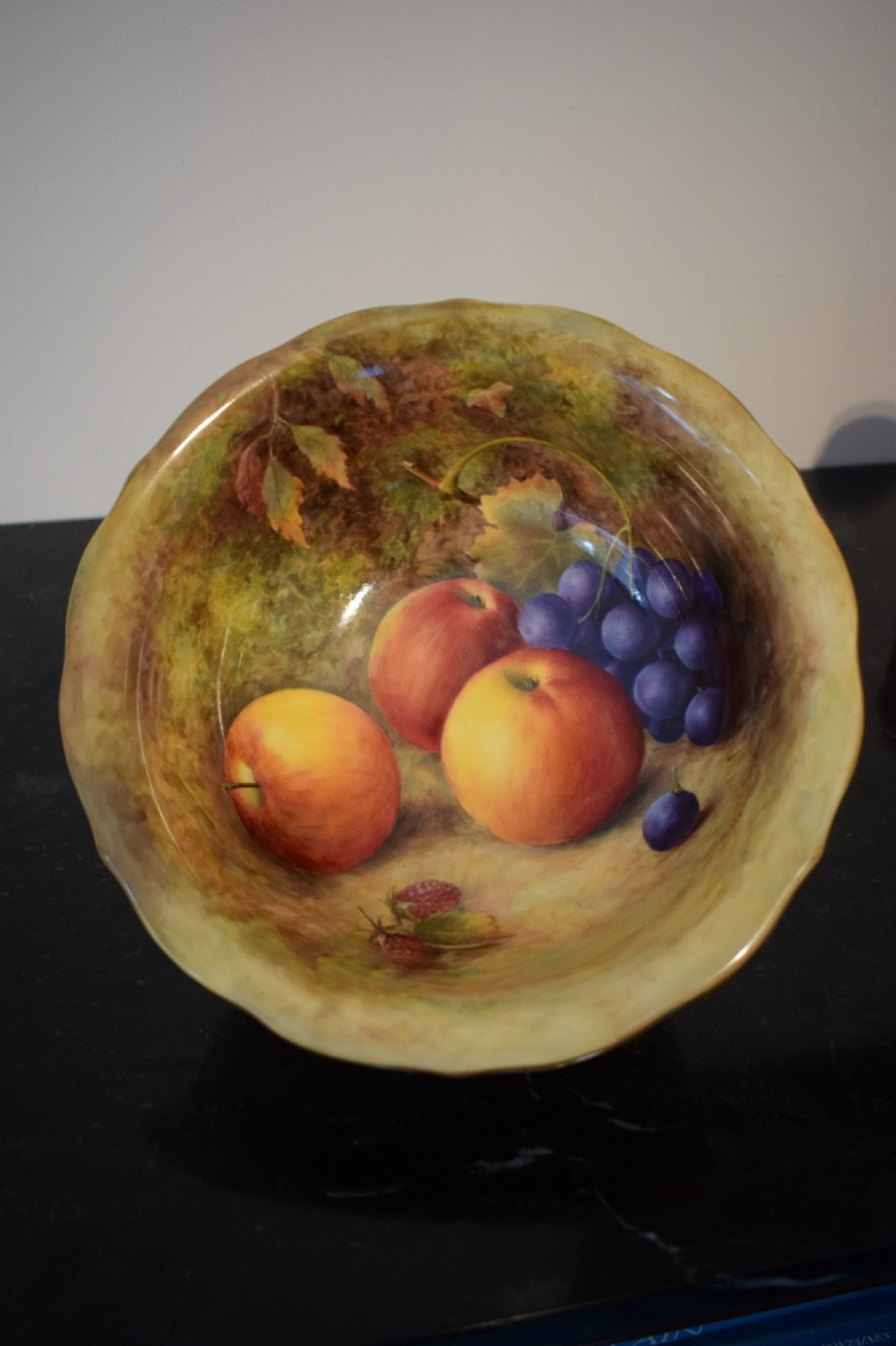 A circular porcelain bowl featuring a detailed hand-painted interior depicting apples, grapes, and berries on a mossy background with a scalloped gold-tone rim.