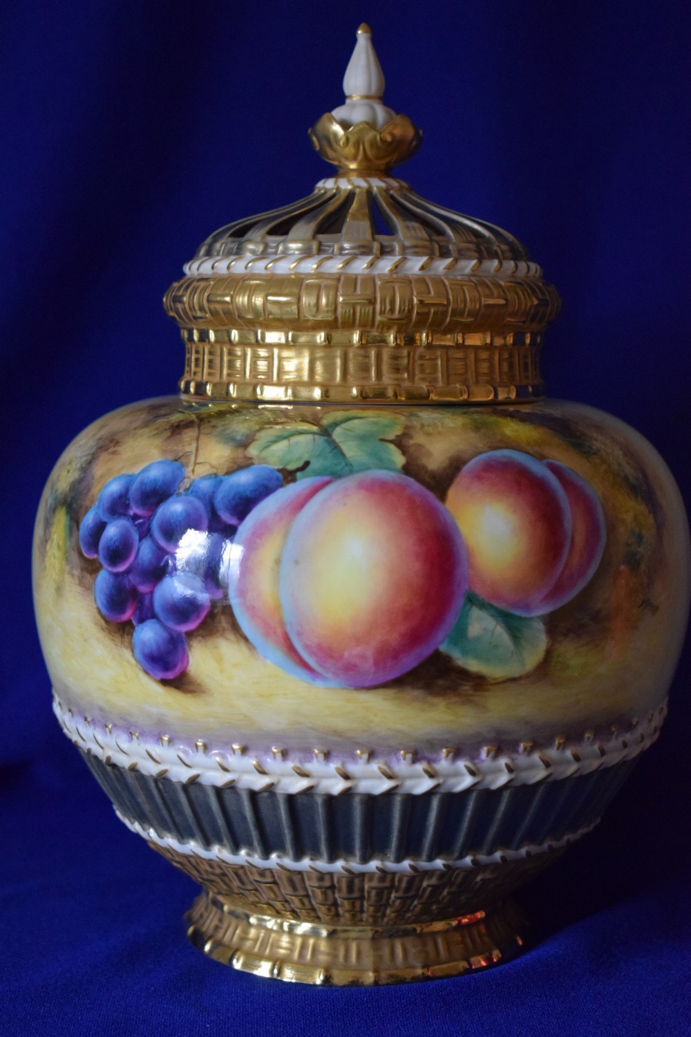 A Royal Worcester three-piece porcelain potpourri vase featuring hand-painted fruit artwork, gilded basketweave lower trim, and a reticulated gold cover.