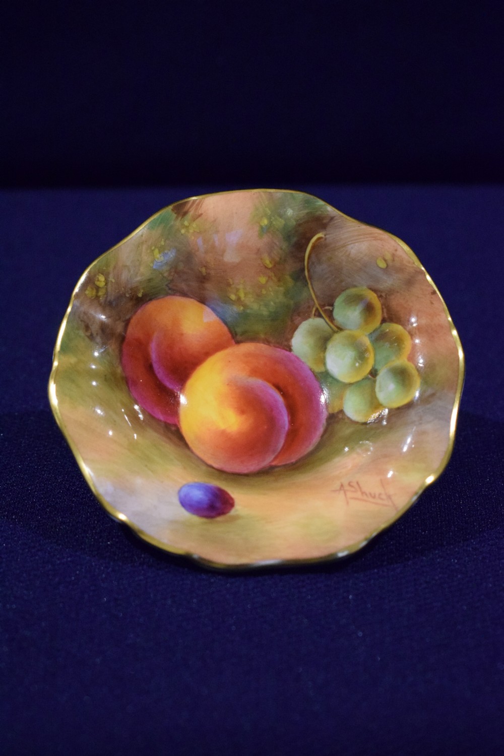 A small circular porcelain dish featuring a scalloped gilded rim and hand-painted peaches and green grapes.