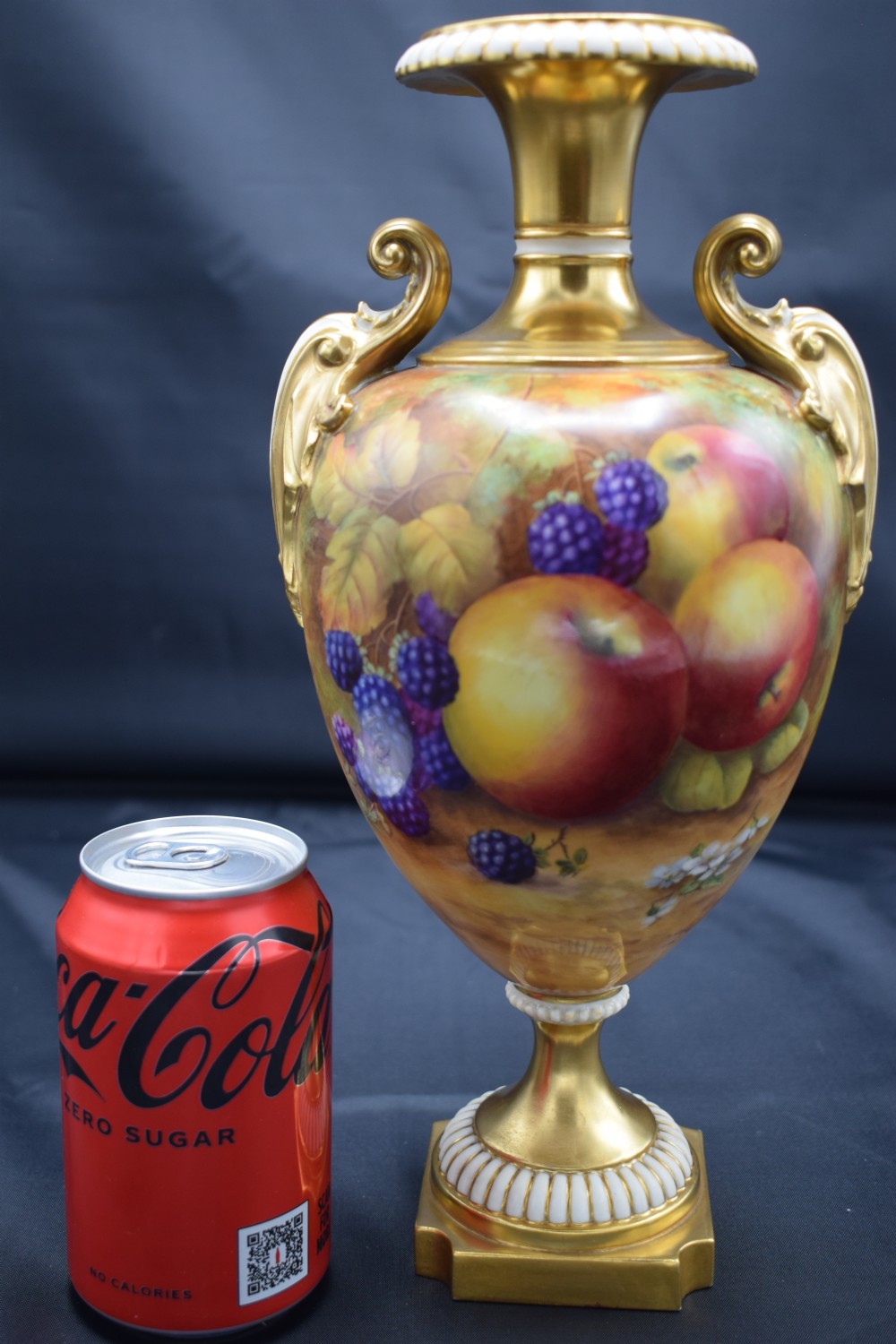 A gilded porcelain urn vase featuring hand-painted apples and blackberries with twin scroll handles and a square pedestal base. View VI