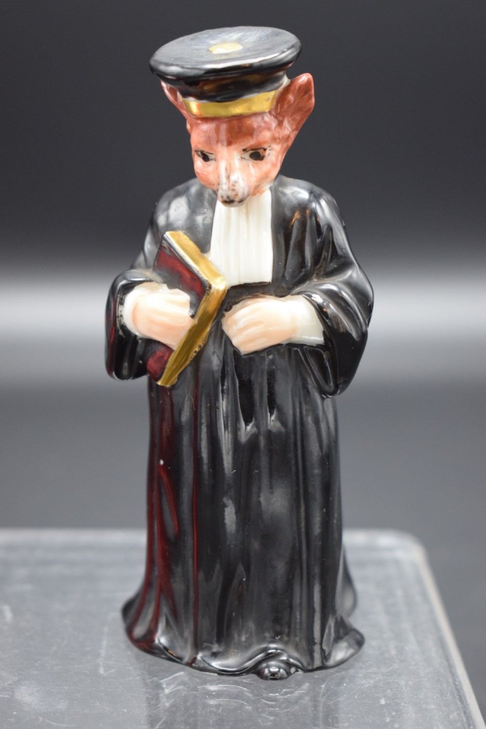 A porcelain candle snuffer shaped as an anthropomorphic fox wearing a black academic gown and mortarboard cap while holding a book.