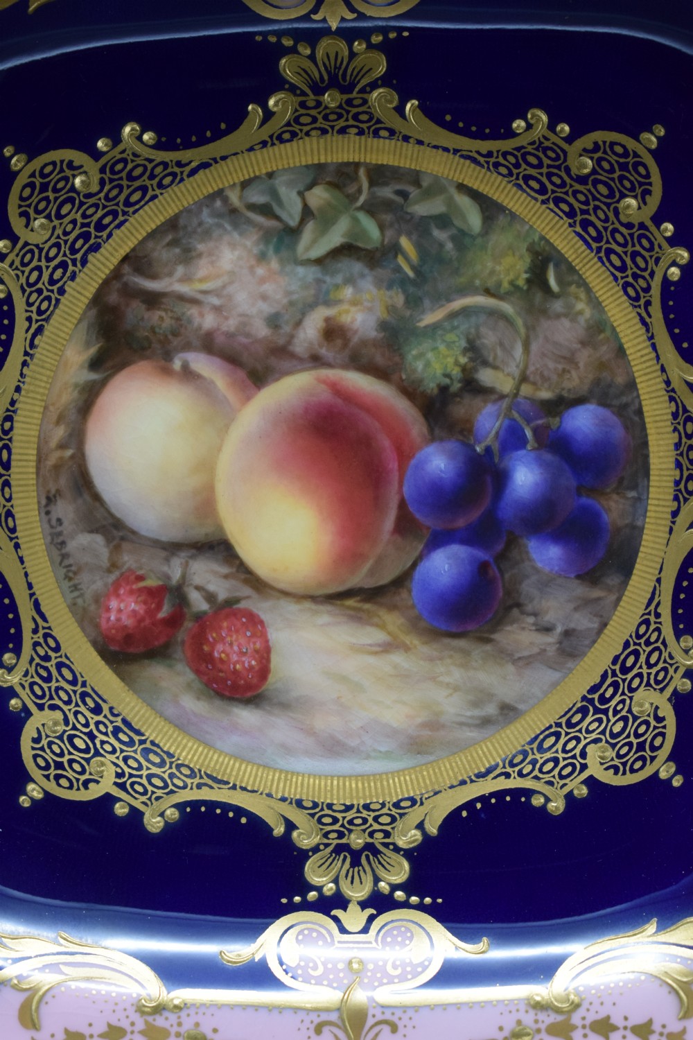 A square scalloped Royal Worcester porcelain dish decorated with a hand-painted fruit panel surrounded by rich cobalt blue, pink, and gilt borders. View II