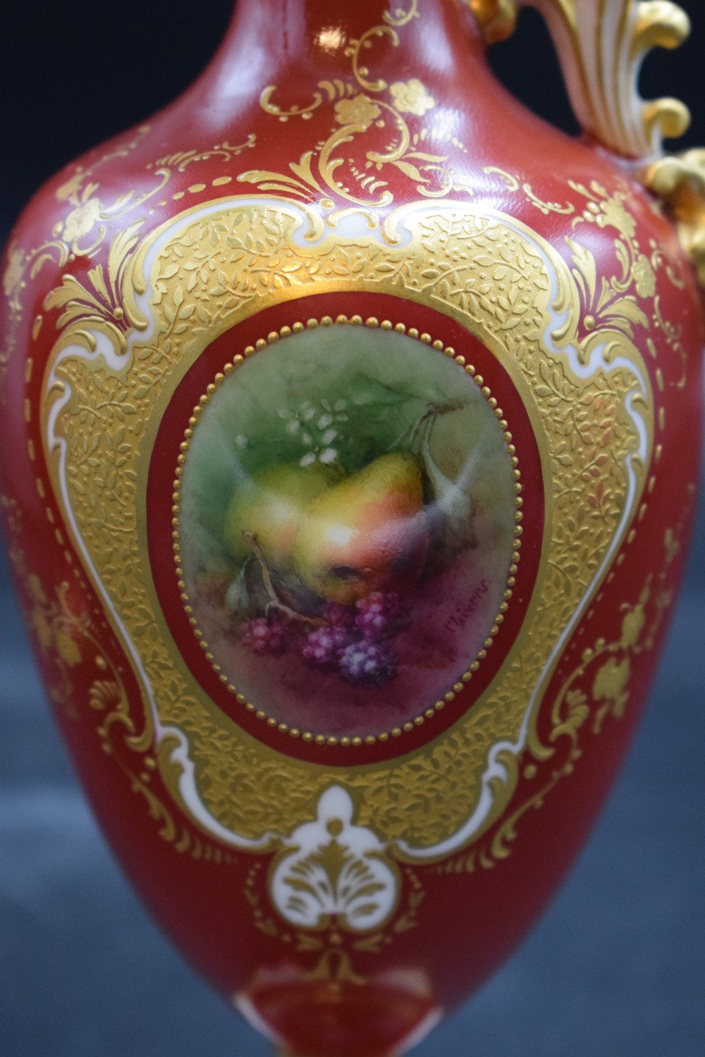 A pair of crimson red porcelain ewers featuring hand-painted fruit vignettes enclosed within gilt scrollwork borders and loop handles on square pedestal bases. View V